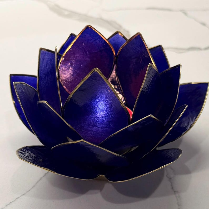 May include: A handcrafted, blue lotus flower candle holder with gold trim. The petals are a deep blue with a metallic sheen, and the interior is a lighter purple. The candle holder is designed to resemble a lotus flower.