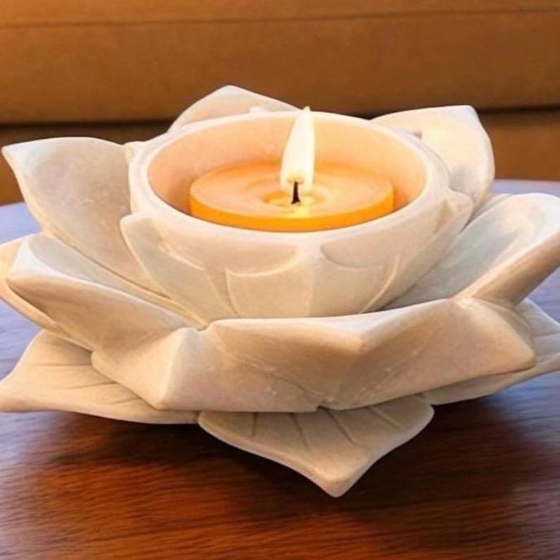 May include: A white lotus flower-shaped candle holder with a lit orange candle. The candle holder is made of a light-colored material and sits on a wooden surface. The flame is bright and the overall aesthetic is zen.