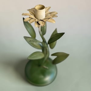 May include: A decorative candle holder shaped like a flower. The candle holder has a cream-colored flower head with a cylindrical candle cup. The stem and leaves are green, and the base is a rounded green shape.