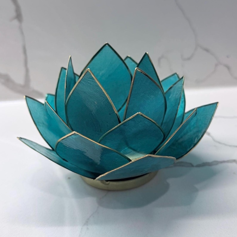 May include: A teal lotus flower candle holder with gold trim. The petals are a translucent teal color, and the edges are outlined in gold. The candle holder sits on a gold base. The overall design is elegant and decorative.