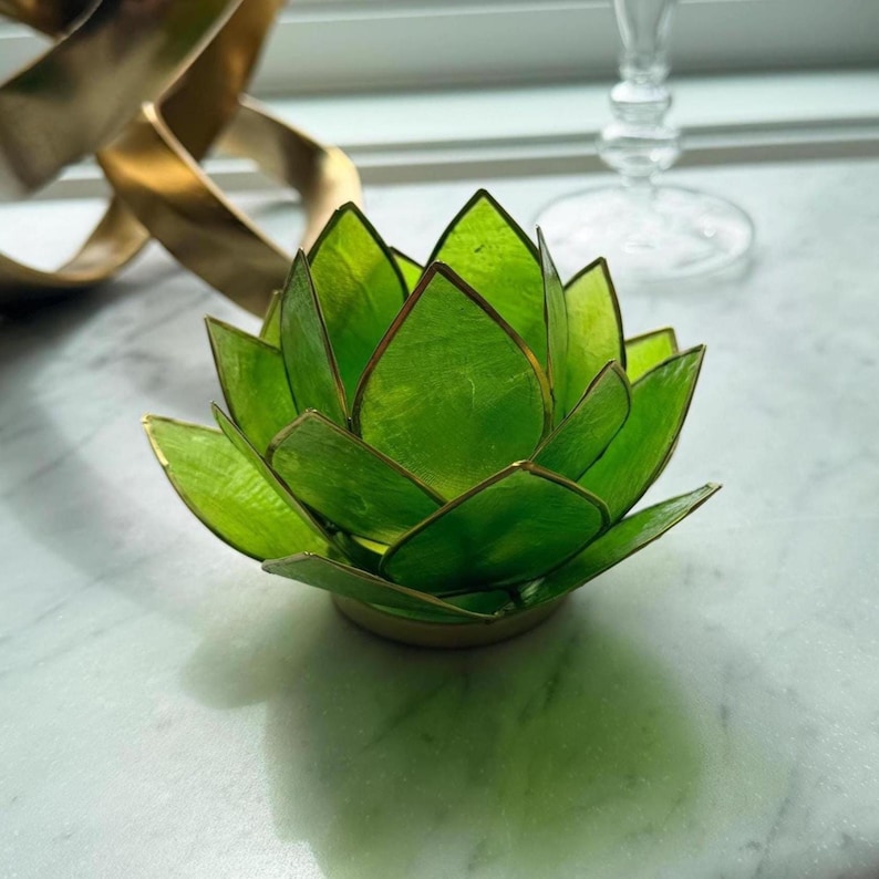 A green lotus flower-shaped candle holder with gold trim. The candle holder is made of translucent green material and sits on a gold base. The petals are layered to create a realistic flower appearance.