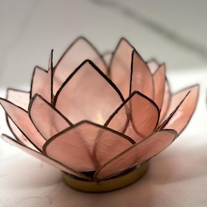 May include: A pink lotus flower candle holder with dark brown outlines. The petals are a soft pink color, and the base is a gold color. The candle holder is designed to hold a small candle.
