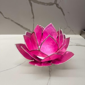 May include: A vibrant pink lotus flower-shaped candle holder. The petals are a bright pink with silver edges. The candle holder is made of metal and sits on a white surface. The overall theme is home decor.