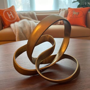 May include: A gold-colored decorative sculpture featuring three interlocking, continuous loops that form a complex, knotted shape. The object has a smooth, metallic finish with subtle variations in light reflection across its curved surfaces.