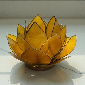 May include: A golden lotus flower-shaped candle holder. The petals are a translucent yellow, and the edges are outlined in a darker gold. The candle holder sits on a round base, casting a shadow.