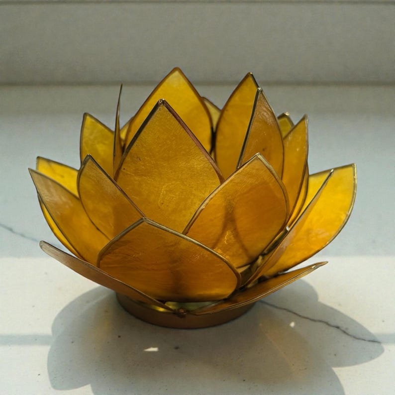 May include: A golden lotus flower-shaped candle holder. The petals are a translucent yellow, and the edges are outlined in a darker gold. The candle holder sits on a round base, casting a shadow.