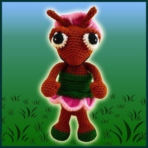 May include: A crocheted ant doll with brown body, green skirt, and pink flower accents. The ant has large black eyes and a pink flower on its head.