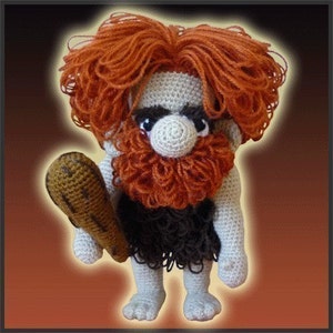 May include: A crocheted plush toy of a caveman holding a club. The caveman has a red beard and hair, a white face, and a brown loincloth.