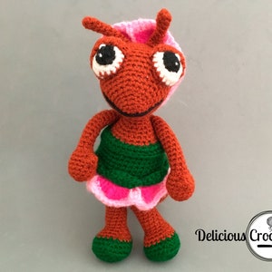 Puede incluir: Una muñeca de hormiga de crochet con un vestido rosa y verde, cuerpo marrón y grandes ojos negros. La muñeca está de pie sobre un fondo gris.