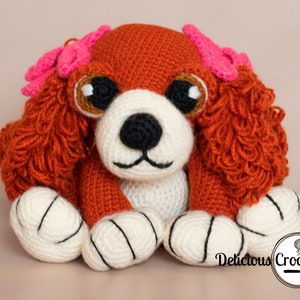 May include: A crocheted stuffed toy dog with brown fur, white paws, and pink bows. The dog has brown eyes and a black nose.