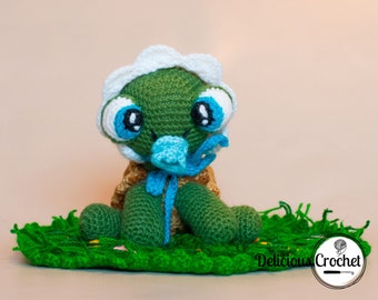 Puede incluir: Una tortuga verde de crochet que lleva un gorro blanco de crochet con ojos azules y un lazo azul. La tortuga está sentada en una manta verde de crochet con una flor amarilla.