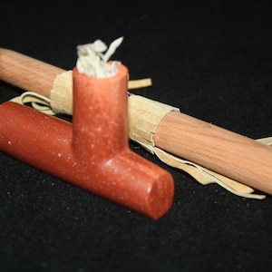 Könnte beinhalten: Eine handgefertigte, rotbraune Steinpfeife mit einem Holzschaft. Die Pfeife hat einen T-förmigen Kopf und einen zylindrischen Schaft. Der Holzschaft ist hellbraun und hat eine Lederwicklung. Die Pfeife ist zum Rauchen bestimmt.