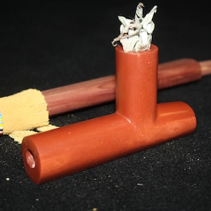 May include: A reddish-brown pipe with a T-shaped design, filled with dried herbs. A wooden stick and a decorated item with colorful beads are in the background. The items are on a black surface.