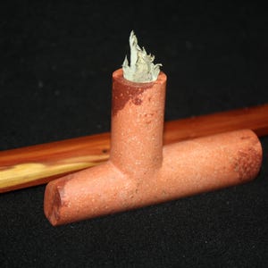 Puede incluir: Una pipa de terracota en forma de T con una superficie texturizada de color marrón rojizo. Una pequeña hoja seca de color claro está colocada dentro del hornillo vertical de la pipa. Una vara de madera lisa de color marrón rojizo está detrás.