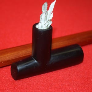 Puede incluir: Una pipa de fumar negra en forma de T con un acabado mate suave. La cámara vertical contiene una pequeña cantidad de material fibroso blanco que sobresale de la abertura superior. La pipa está colocada horizontalmente sobre un fondo rojo.