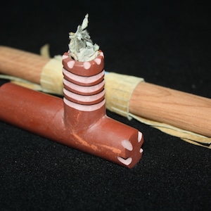 Könnte beinhalten: Eine rotbraune, geschnitzte Steinpfeife mit zylindrischem Kopf und Stiel. Die Pfeife ist mit einer hellfarbigen Substanz gefüllt. Ein Holzstab mit beiger Umwicklung liegt hinter der Pfeife.