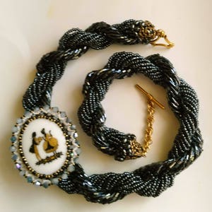 May include: A black beaded necklace with a silver and white cameo pendant featuring two women in a vintage style. The necklace has a gold clasp and a gold chain extender.