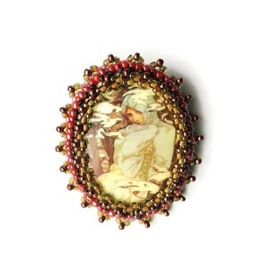 May include: Oval brooch featuring a vintage illustration of a person in winter attire. The image is framed by a beaded border in shades of red, brown, and gold. The brooch is set against a white background.