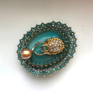 May include: A teal blue and gold beaded brooch with a jeweled beetle design. The brooch features a large, round, teal blue stone with a gold beetle design in the center. The beetle has a green gem for an eye and is covered in clear rhinestones. The brooch is surrounded by a beaded border in shades of teal blue and gold.