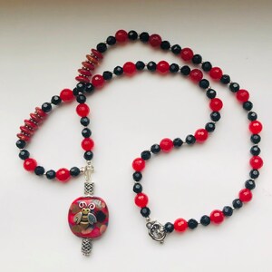 May include: A red and black beaded necklace with a silver charm featuring a bee design. The charm is made of glass and has a multi-colored pattern.