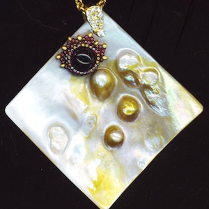 May include: A square-shaped pendant made of iridescent mother-of-pearl with a gold chain and a dark red and black beaded embellishment.