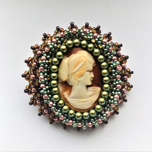 May include: A cameo brooch with a white profile of a woman set in a brown shell. The cameo is surrounded by a beaded border with green, pink, and brown beads.