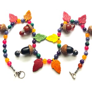May include: A colorful necklace with acorn and leaf charms. The necklace is made with beads in shades of orange, yellow, green, purple, and pink. The acorns are brown and black, and the leaves are red, orange, yellow, and green.