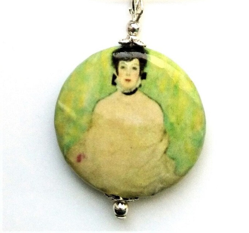 May include: A round pendant necklace featuring a painting of a woman in a white dress with a green background. The pendant is made of ceramic and has a silver chain.