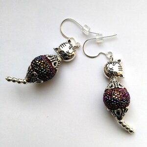 May include: A pair of silver cat earrings with iridescent purple and gold beads. The cat faces are detailed and the beads are covered in tiny sparkles.