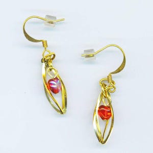 May include: A pair of gold-colored earrings. Each earring features a red heart-shaped bead suspended within a gold-toned, leaf-shaped frame. The earrings have a hook-style closure with a clear, plastic backing.