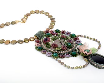 May include: A beaded necklace with a large, ornate pendant featuring a green, pink, and purple floral design. The pendant is surrounded by a circle of green stones and the necklace is made of brown pearls.