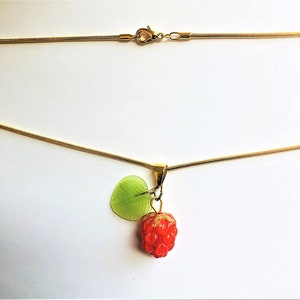 May include: A gold-colored necklace with a pendant featuring a red raspberry and a green leaf. The necklace has a delicate chain and a lobster clasp closure. The pendant is detailed and realistic, with a shiny finish.