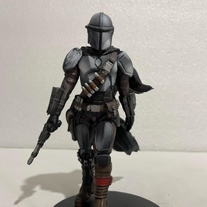 May include: A grey and black action figure of a Mandalorian with a blaster rifle, wearing a helmet and armour, standing on a black circular base.