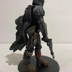 May include: A black and grey action figure of a character wearing a helmet and armour, holding a blaster pistol. The figure is standing on a black circular base.