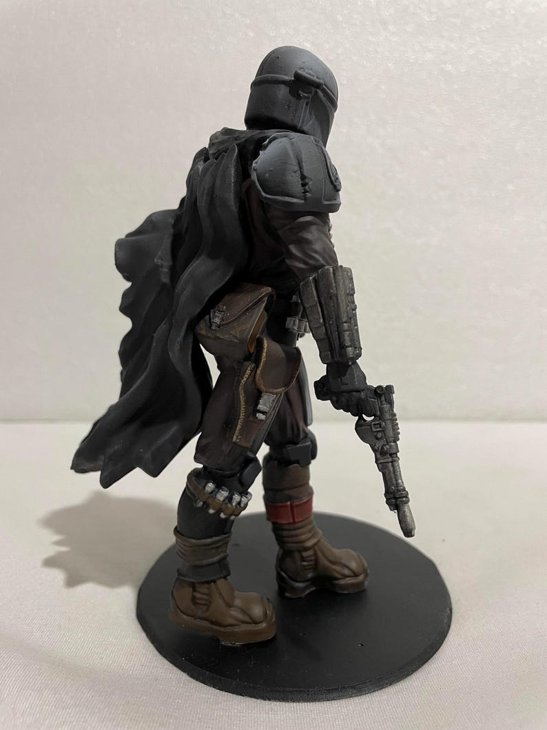 May include: A black and grey action figure of a character wearing a helmet and armour, holding a blaster pistol. The figure is standing on a black circular base.
