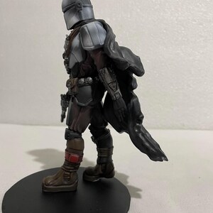 May include: A grey and black action figure of a character wearing a helmet and a cape. The figure is standing on a black circular base.