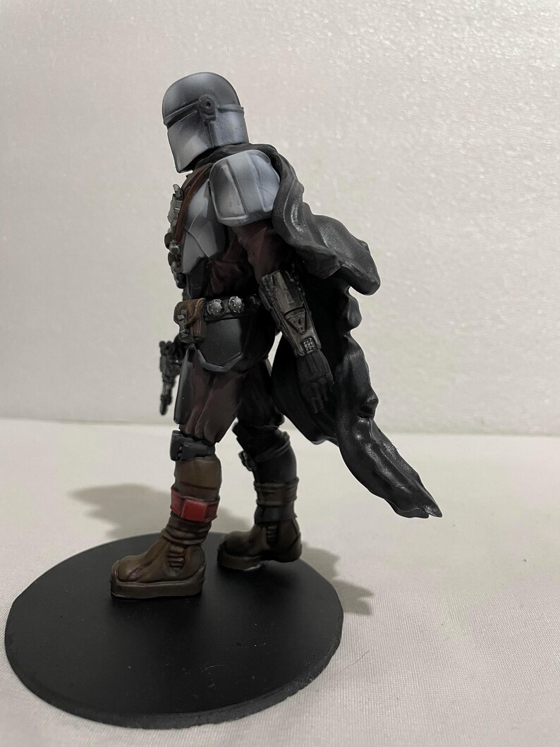 May include: A grey and black action figure of a character wearing a helmet and a cape. The figure is standing on a black circular base.