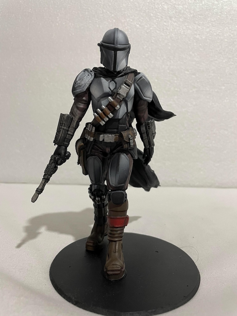 May include: A grey and black action figure of a Mandalorian with a blaster rifle, wearing a helmet and armour, standing on a black circular base.