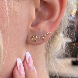 May include: A gold-toned metal ear climber earring featuring the name Raegan written in a cursive script font. The earring follows the curve of the earlobe, with the letters connected in a single, continuous line of polished metal.