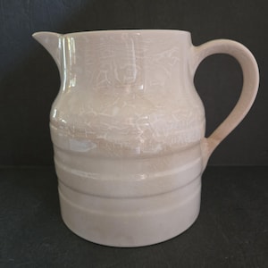May include: A cream-colored ceramic pitcher with a spout and a curved handle. The pitcher has a textured, crackled surface and horizontal bands around the base. This decorative pitcher is suitable for serving beverages.