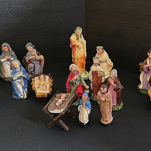 May include: A collection of hand-painted nativity figurines including multiple figures of adults in robes, several infants in cradles, and a small wooden manger. The set features various colors such as blue, red, yellow, and brown on the clothing.
