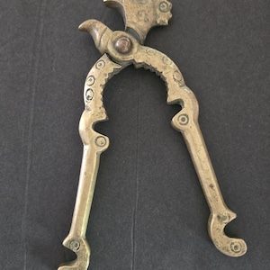 May include: Antique brass nutcracker with a hammer-like head and ornate, curved handles. The nutcracker features a toothed jaw for gripping nuts and decorative circular accents. The brass has a warm, aged patina.