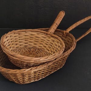 May include: Two woven wicker baskets. One is oval-shaped with a handle, and the other is round with a handle. Both are made of light brown wicker and are empty. The baskets are likely for serving bread or other items.