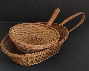 May include: Two woven wicker baskets. One is oval-shaped with a handle, and the other is round with a handle. Both are made of light brown wicker and are empty. The baskets are likely for serving bread or other items.