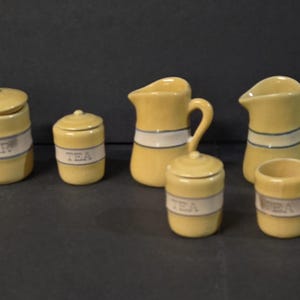 May include: A collection of yellow ceramic kitchen canisters and pitchers. The canisters are labeled "FLOUR", "SUGAR", and "TEA" with a white and blue band. The pitchers have a similar design. The set is arranged on a dark surface.