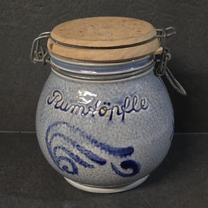 May include: A blue ceramic jar with a wooden lid and metal clasp. The jar has a textured surface and is decorated with blue swirls and the word "Rumtöpfe". A decorative kitchen storage container.