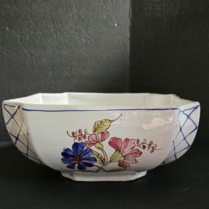 May include: A white ceramic bowl with a scalloped rim and a footed base. The exterior features a hand-painted floral motif in blue, pink, and green, complemented by a blue cross-hatch pattern along the sides and a thin blue line near the rim.