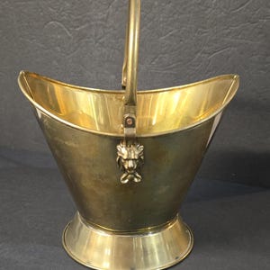 May include: A vintage brass coal scuttle with a handle. The scuttle has a flared, boat-shaped body and a round base. A lion's head adorns the front. The handle is attached to the body with hinges. The scuttle is made of shiny brass.
