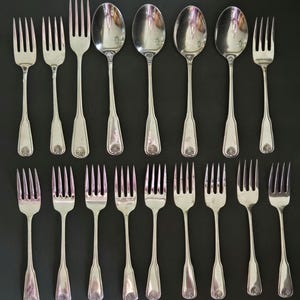 May include: A collection of sixteen silver-toned metal utensils arranged in two rows. The set includes four spoons and twelve forks, each featuring a polished finish and a decorative shell-like motif embossed at the base of the handle.