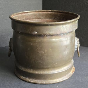 May include: A cylindrical brass vessel with a weathered, dark patina finish. The container features two decorative side handles shaped like lion heads, each with a small attached ring. The rim is smooth and circular, with a wide, open top.